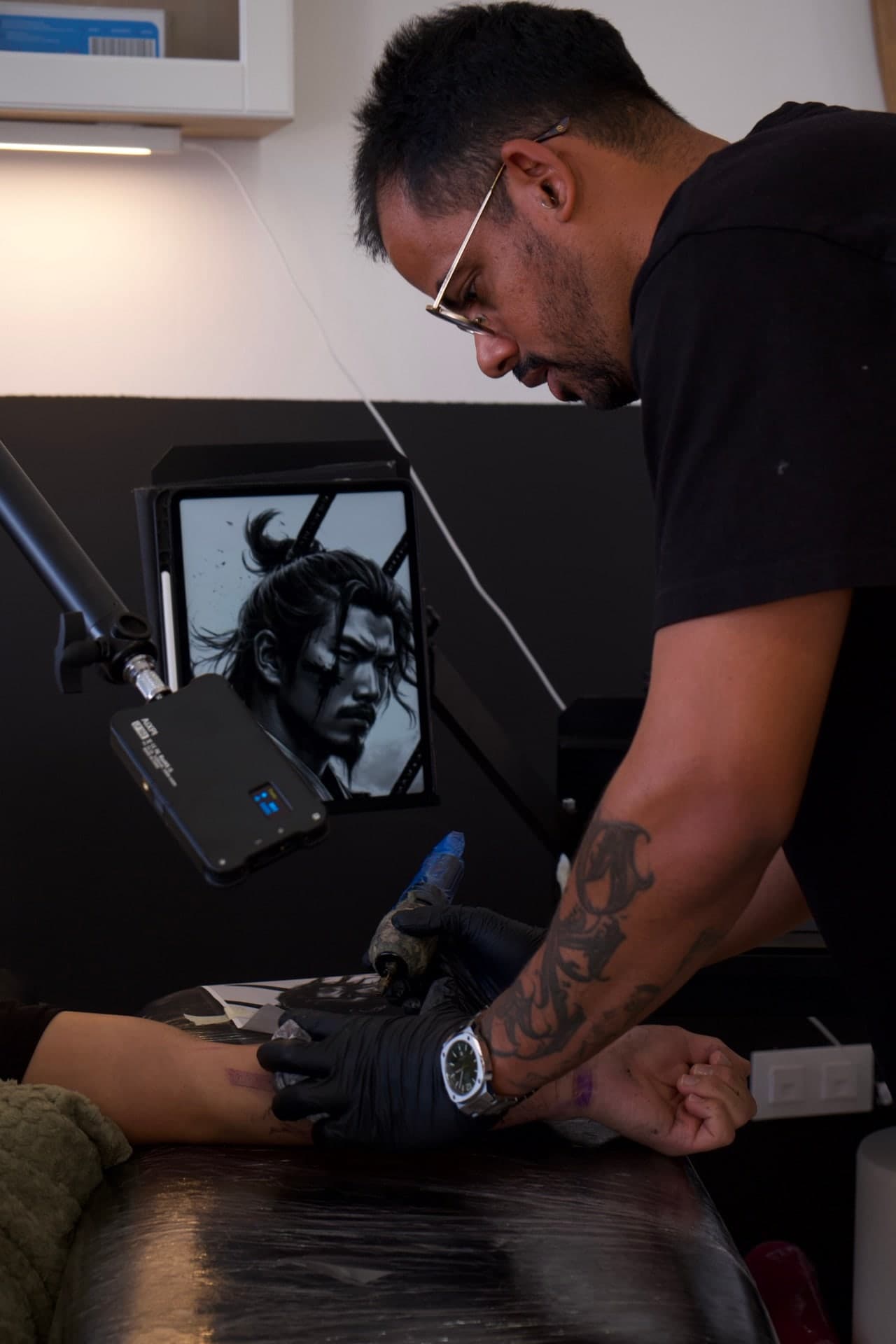 Jubi Pereira, in glasses and black gloves, tattooing a client’s arm at the studio.