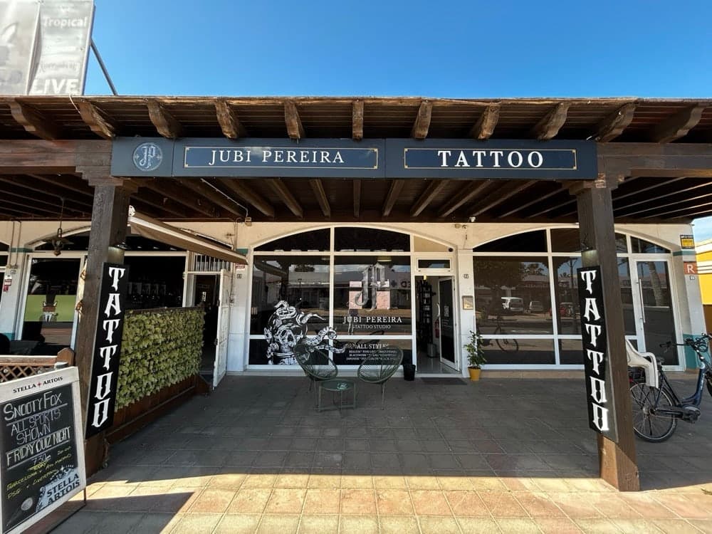 The studio front on a sunny day: a wooden porch with a sign reading Jubi Pereira Tattoo.