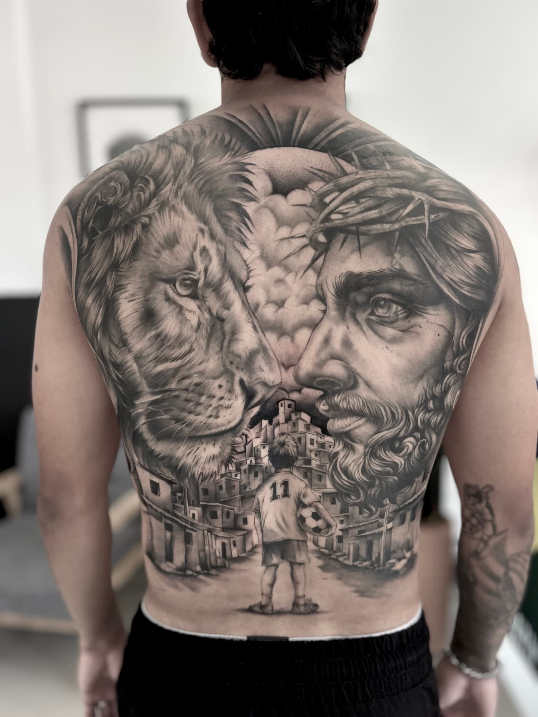 Full back piece in black and grey: a lion and Christ crowned with thorns face each other above a favela, where a boy in a number 11 shirt holds a football.